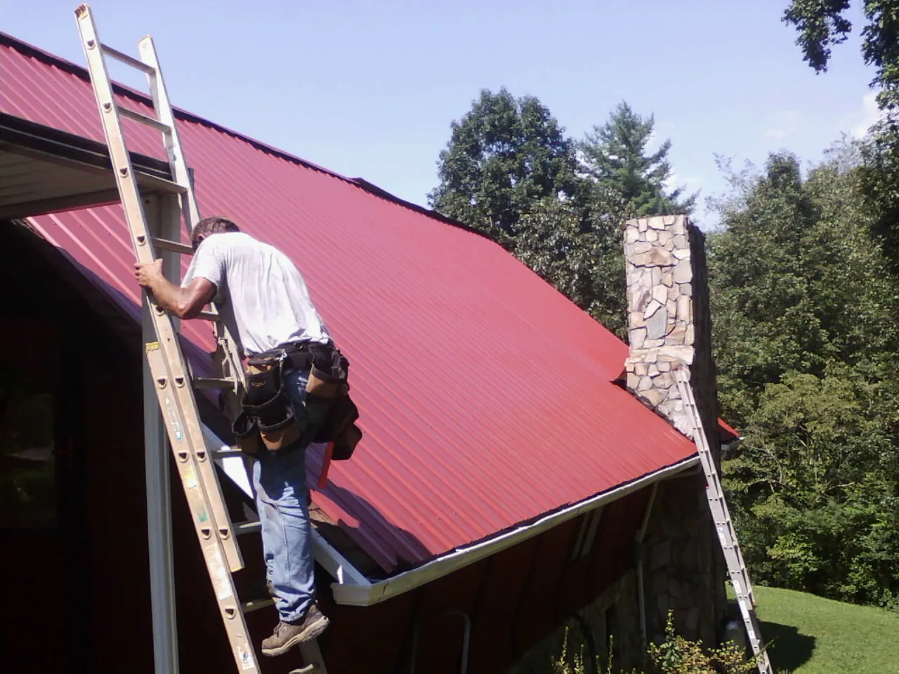 Roofer on a ladder beside a red metal roof and stone chimney — Soffit & Fascia Repair by Roofing Brothers in Alto